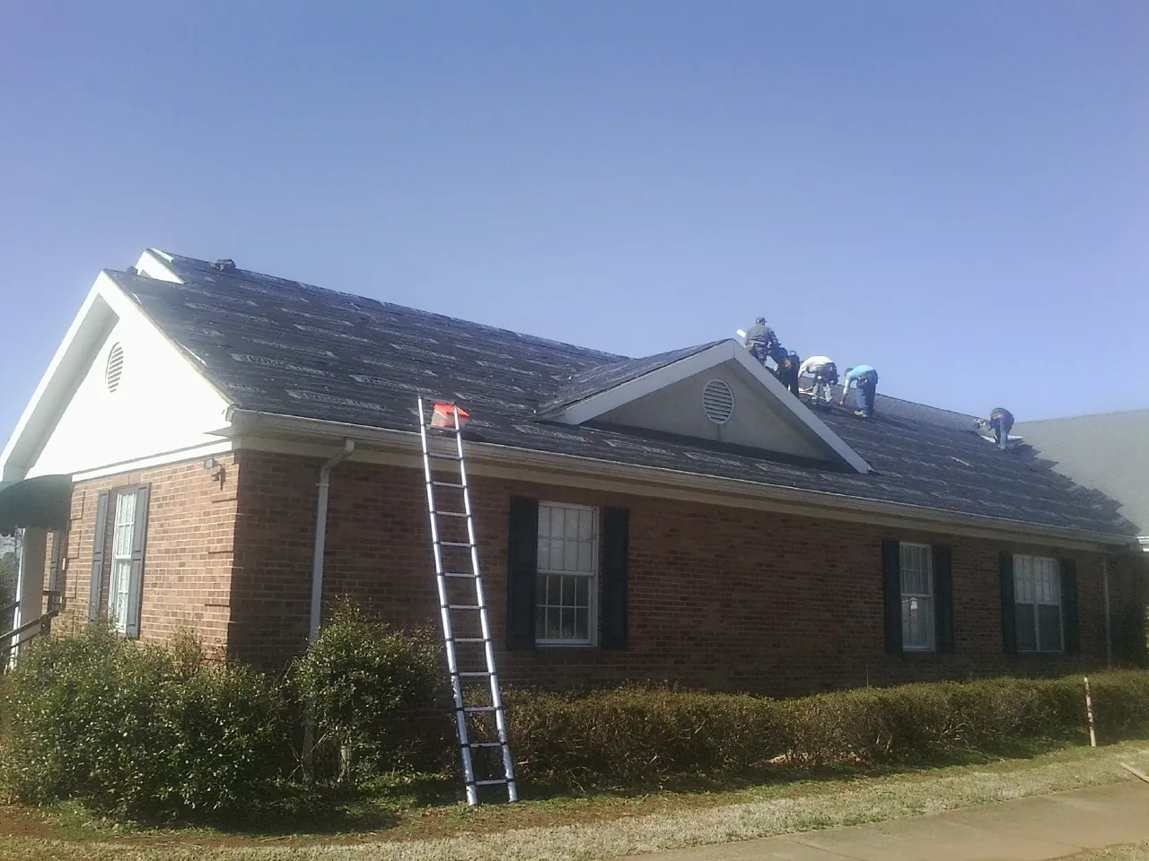 Crew tearing shingles off a brick ranch roof with a ladder set — Emergency Roof Repair work by our crew in Manley