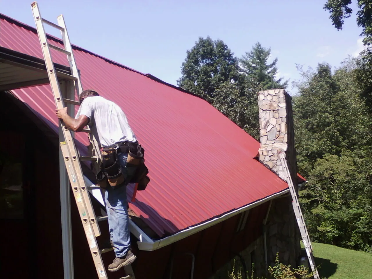 Roofer on a ladder beside a red metal roof and stone chimney — Soffit & Fascia Repair by Roofing Brothers in Manley